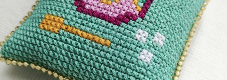 Close up of a small cross stitched pillow featuring a sugar bowl, spoon and sugar cubes